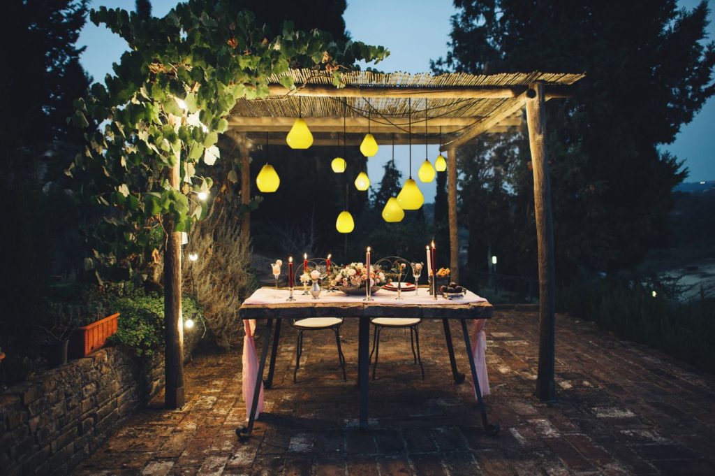 میز عذاخوری همراه با لامپ های آویزان قرار گرفته روی سقف بالای آن واقع در یک باغ